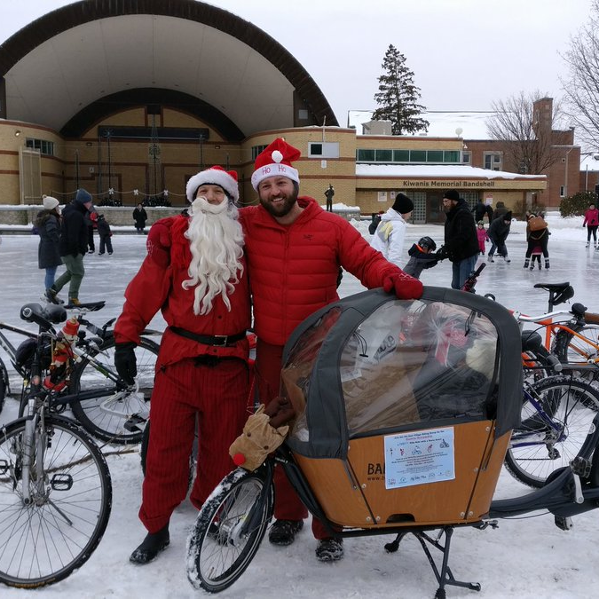 Ben and a Santa friend stand next to the Babboe bakfiets in front of a skating rink, beyond which is a large outdoor bandshell.