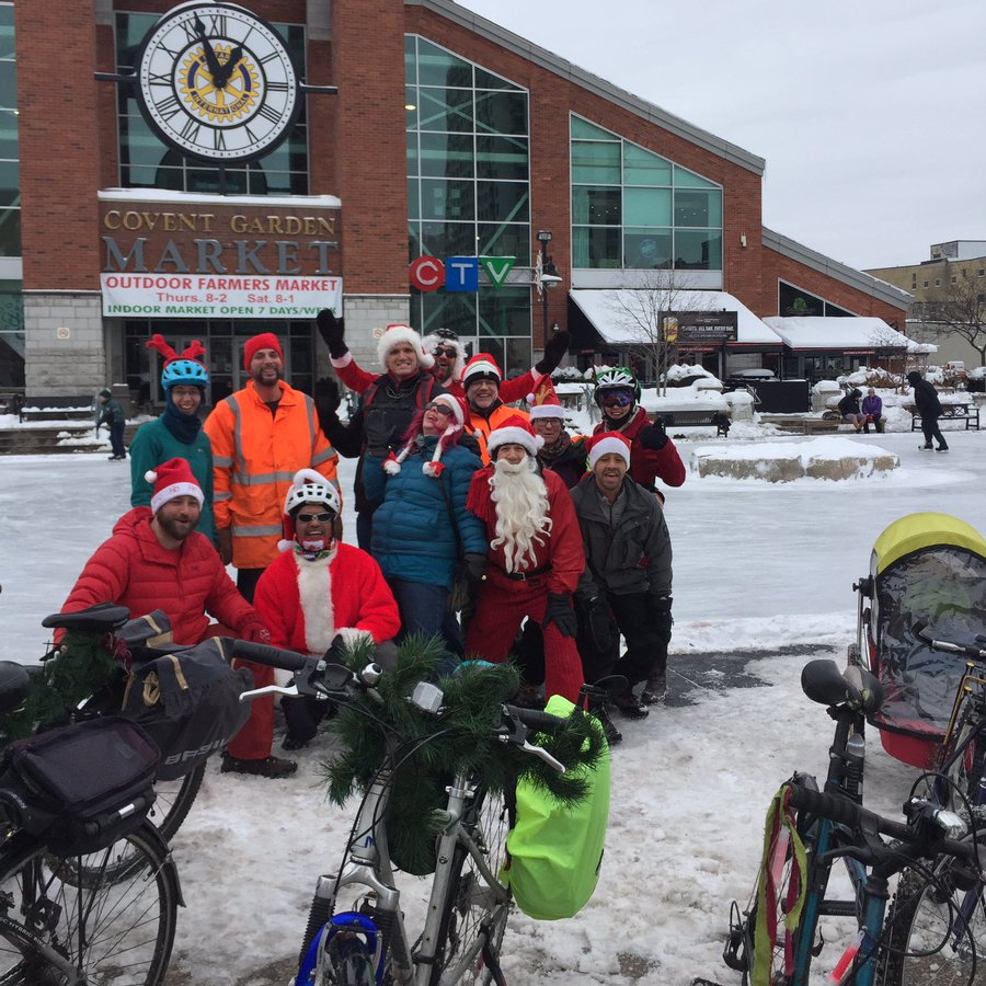 Cyclists dressed a bit like santa smile at the camera, victorious, in front of a skating rink, beyond which is the Covent Garden Market, London, Ontario.