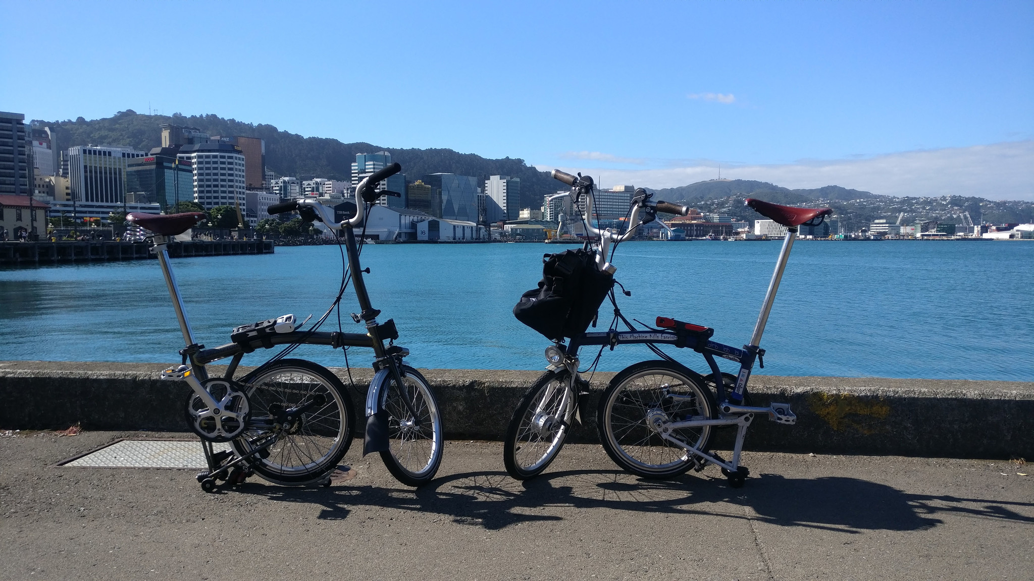 Two Brompton bicycles in front of a waterfront skyline.