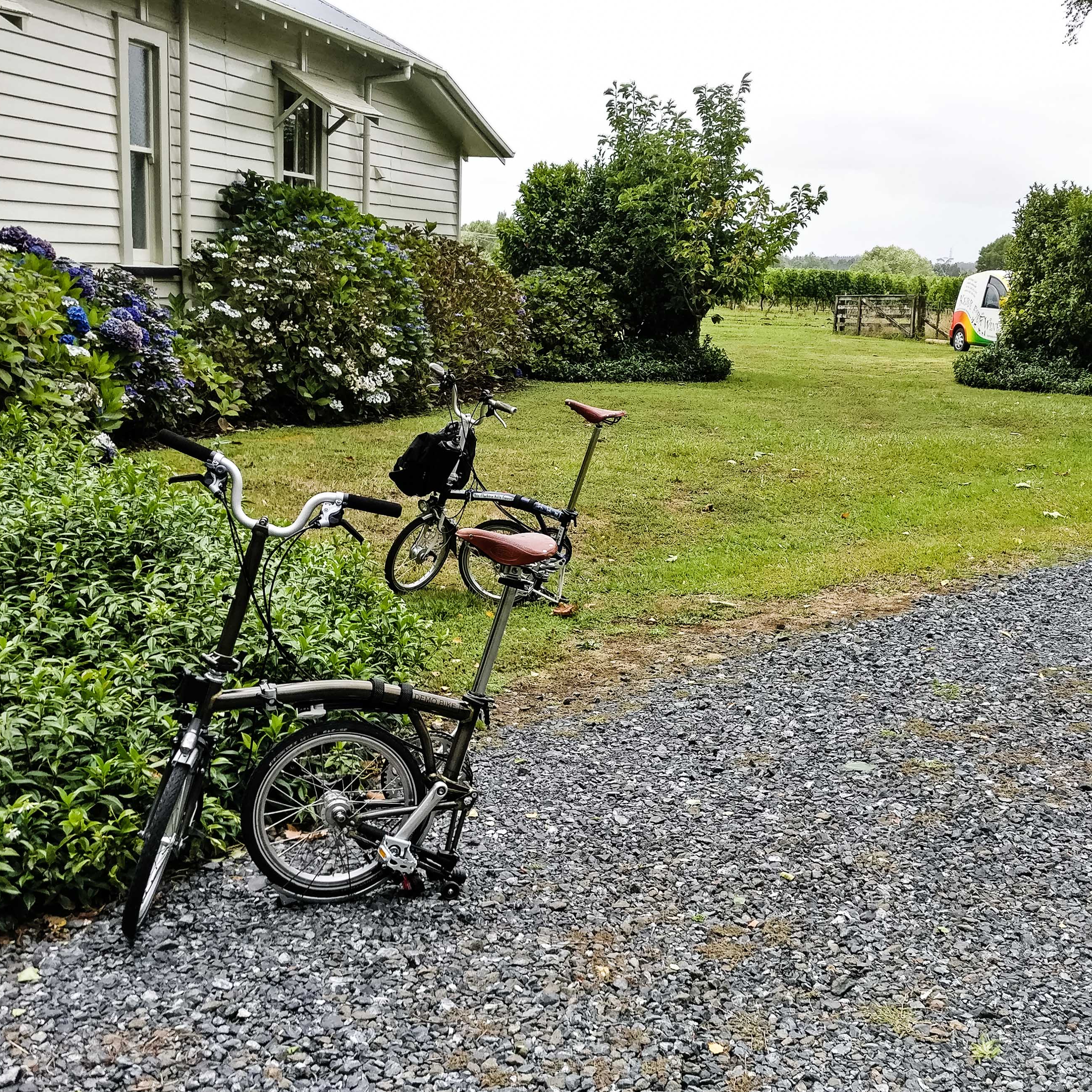 Two Bromptons on a gravel drive by a house surrounded by flowering bushes.