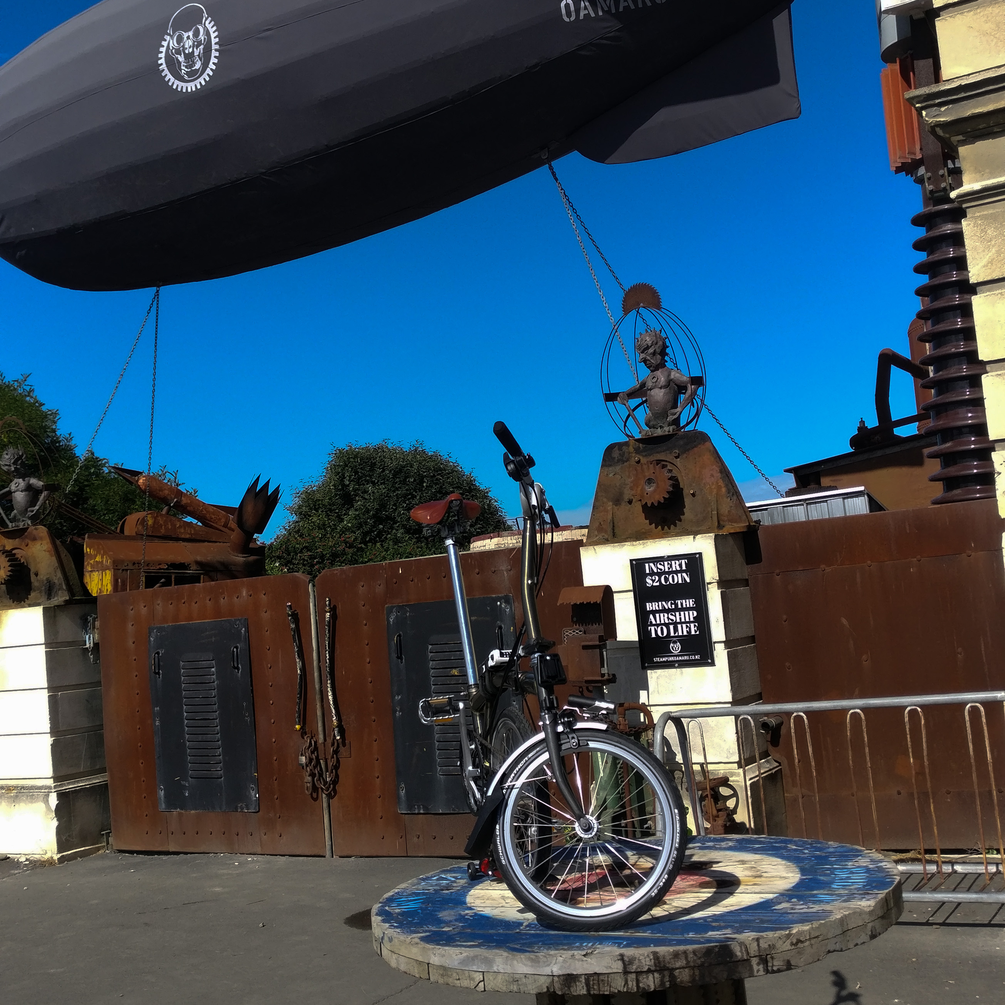 A folded Brompton on a wooden circular table or stand under a mini blimp with a skull on it.