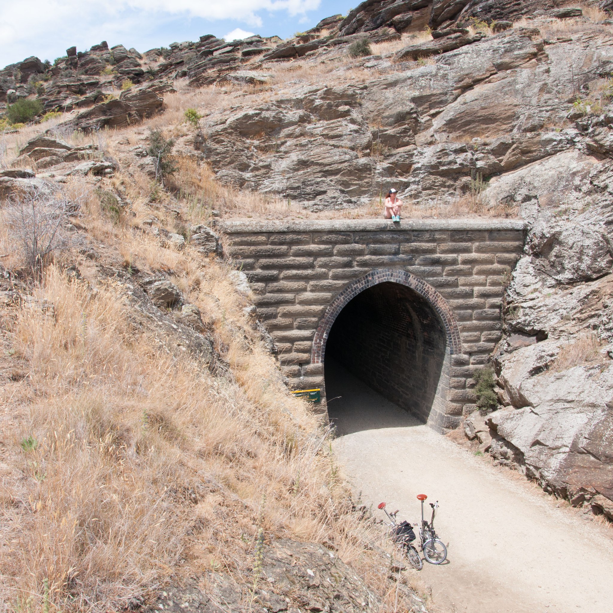 Caroline sits on top of a tunnel entrance in the distance, desert all around, and her Brompton at the entrance of the tunnel.