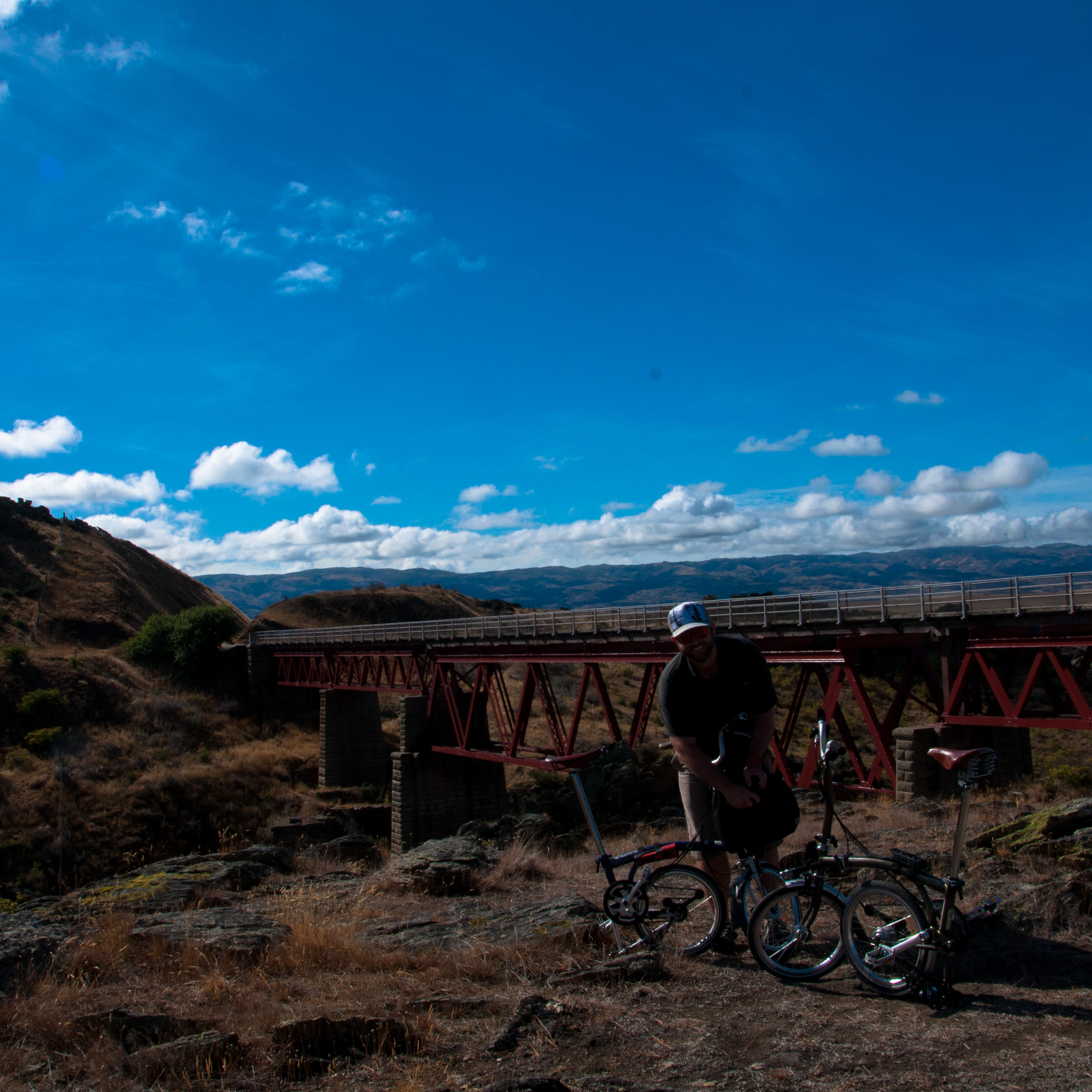 Ben folds his Brompton in front of a more lush vista, a rail bridge and blue skies beyond.