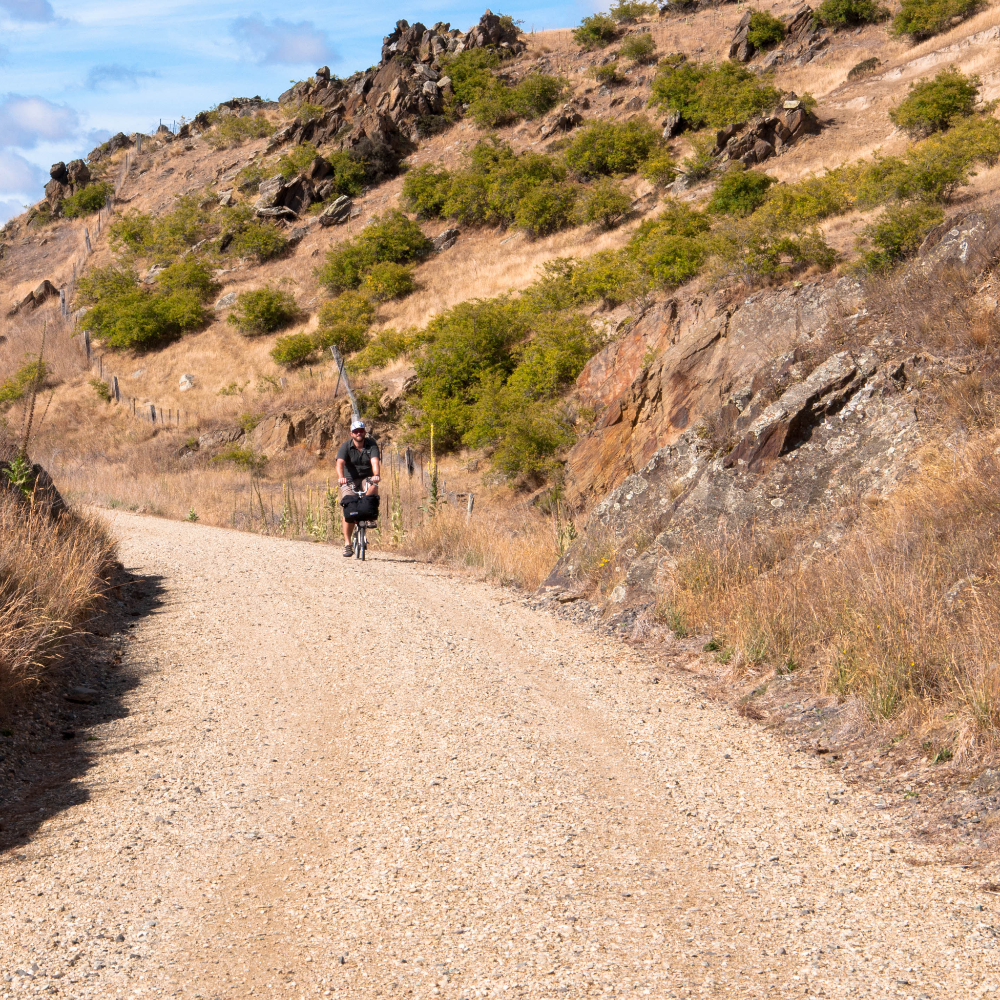 Ben rides his Brompton towards us in the distance along a trail, hilly desert surrounds.