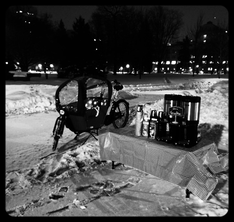 The Babboe city sits along a snowy path in the dark next to a table with coffee and cups.