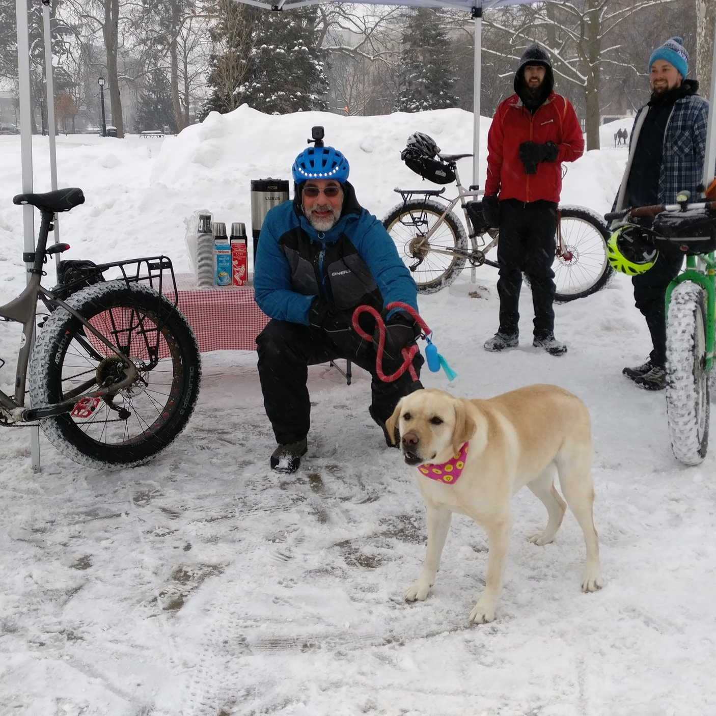 A winter cyclist kneels down in front of the coffee station, facing the camera, with a dog.