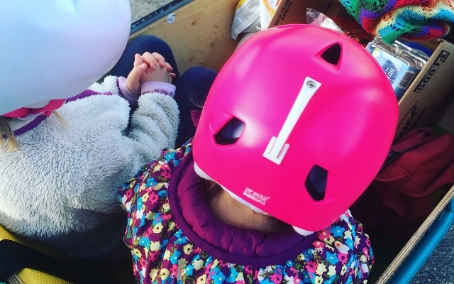 Two helmeted kids from behind in their box bike.