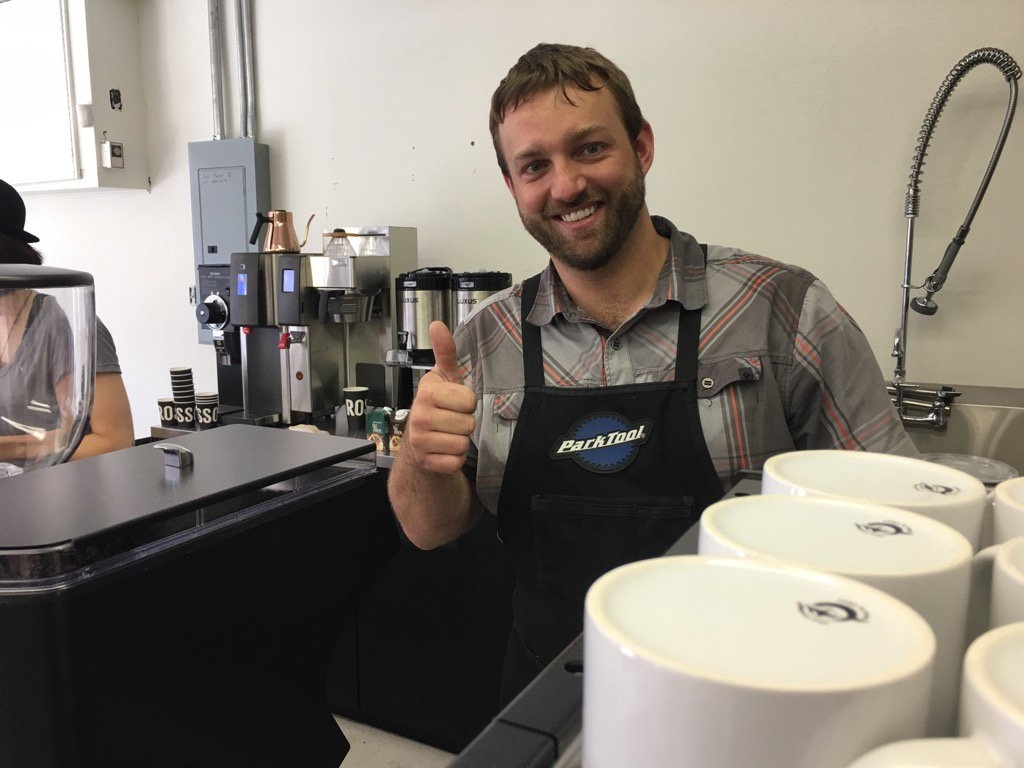 Ben smiles and gives a thumbs up from behind the espresso bar, he wears a Park Tools apron.