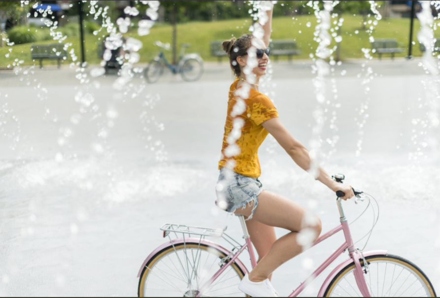 Water splashes directly in front of the camera, beyond which a woman rides a pink step through bicycle and waves, smiling.