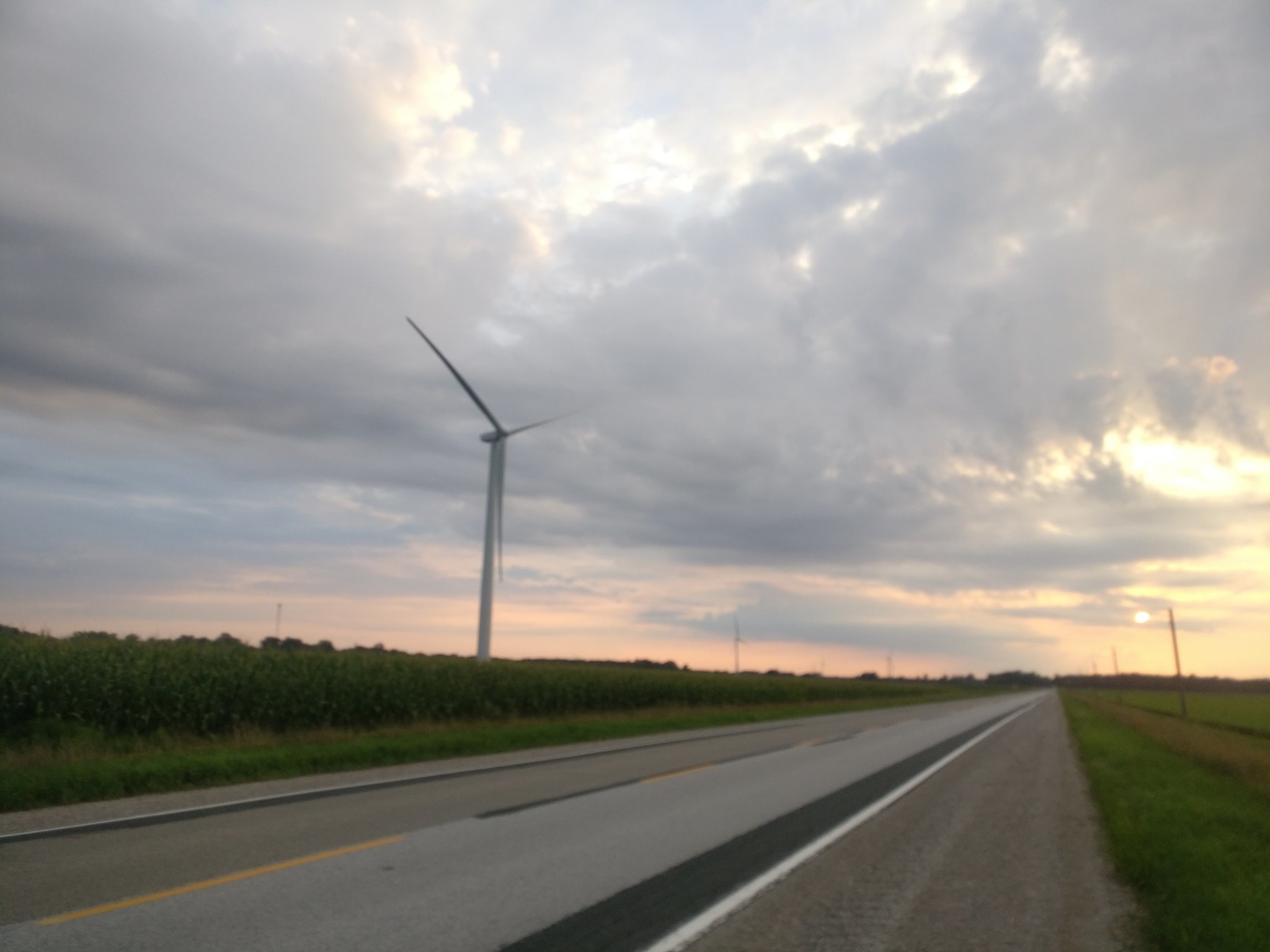 The sun starts to set on a county road, windmills beyond.