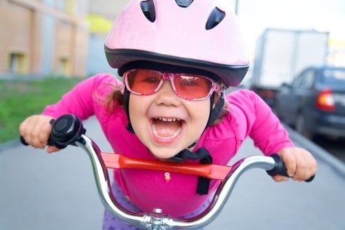 A child leans over her bicycle handlebars at the camera with a huge grin.