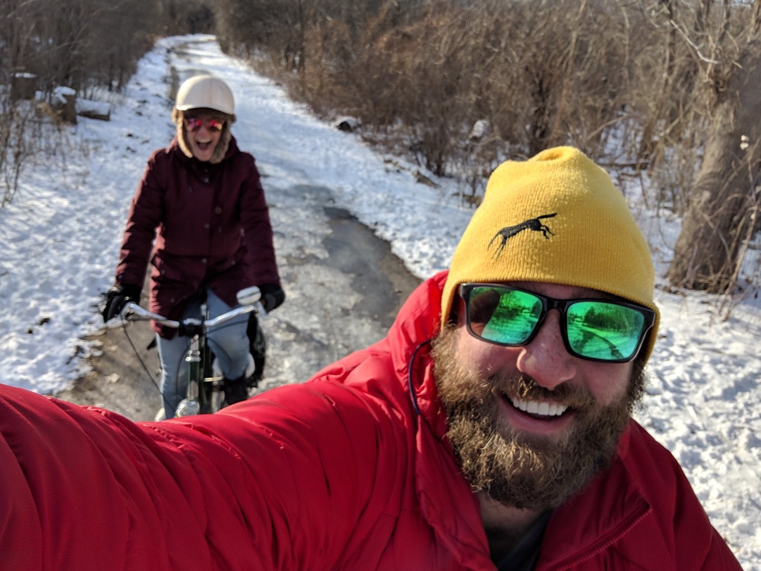 Ben takes a selfie over his shoulder as he rides a snowy winter path; a woman smiles on her bike behind him