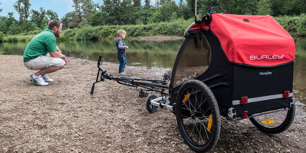 A father kneels next to a toddler by a river, their Burley Bee trailer in the foreground.