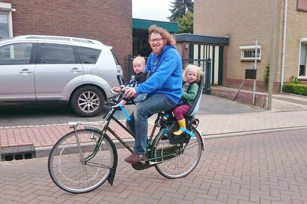 A dad rides a regular bike with a child in a front seat and a rear.