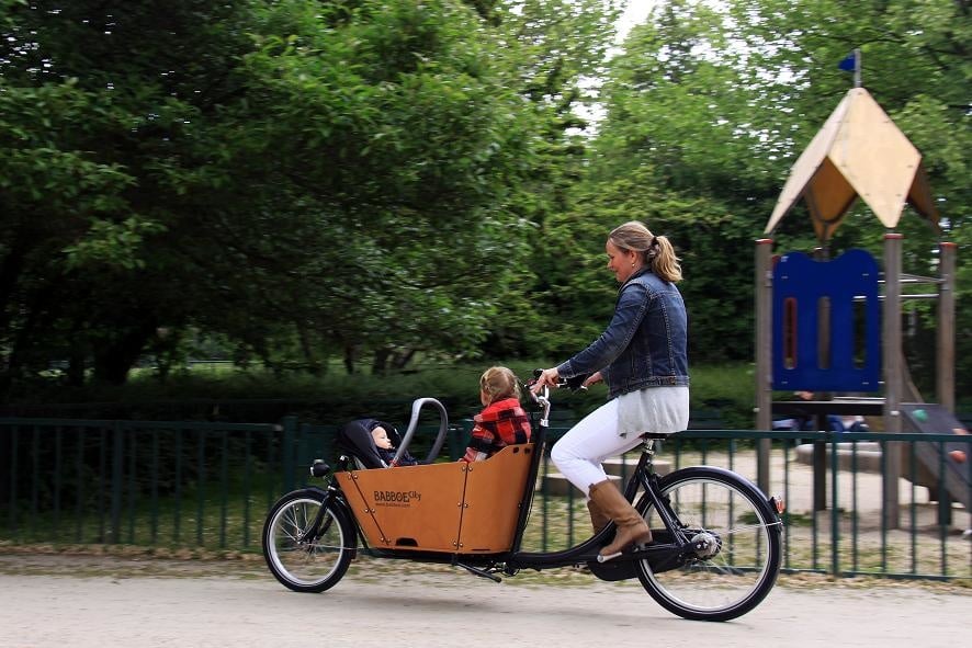 A mom pedals a Babboe City bakfiets with a child in the front box, a park is beyond them.