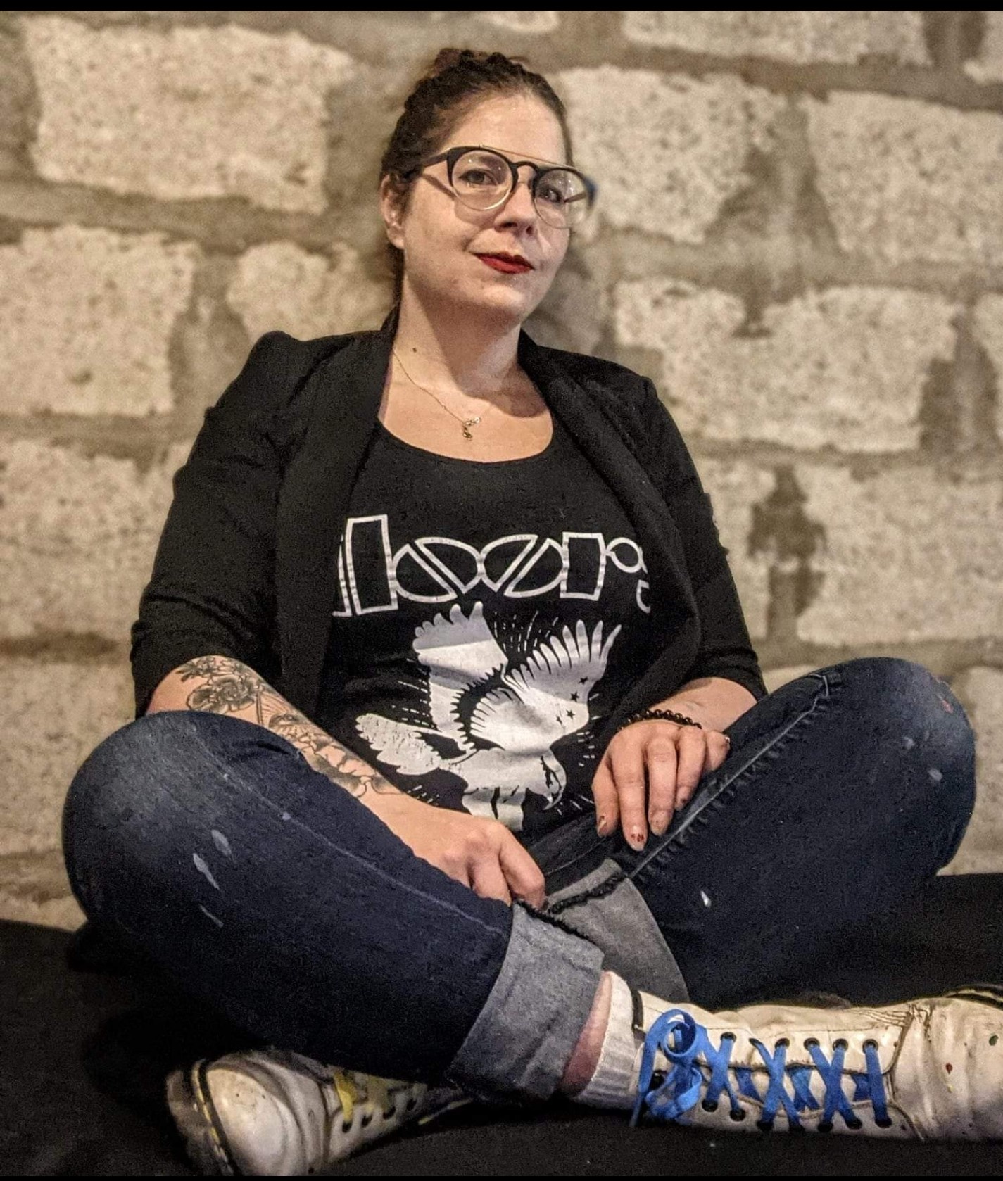 Alicia sits crossed legged against a brick wall and half-smiles at the camera. She wears Docs and a denim jacket over a t-shirt that says "Doors". 