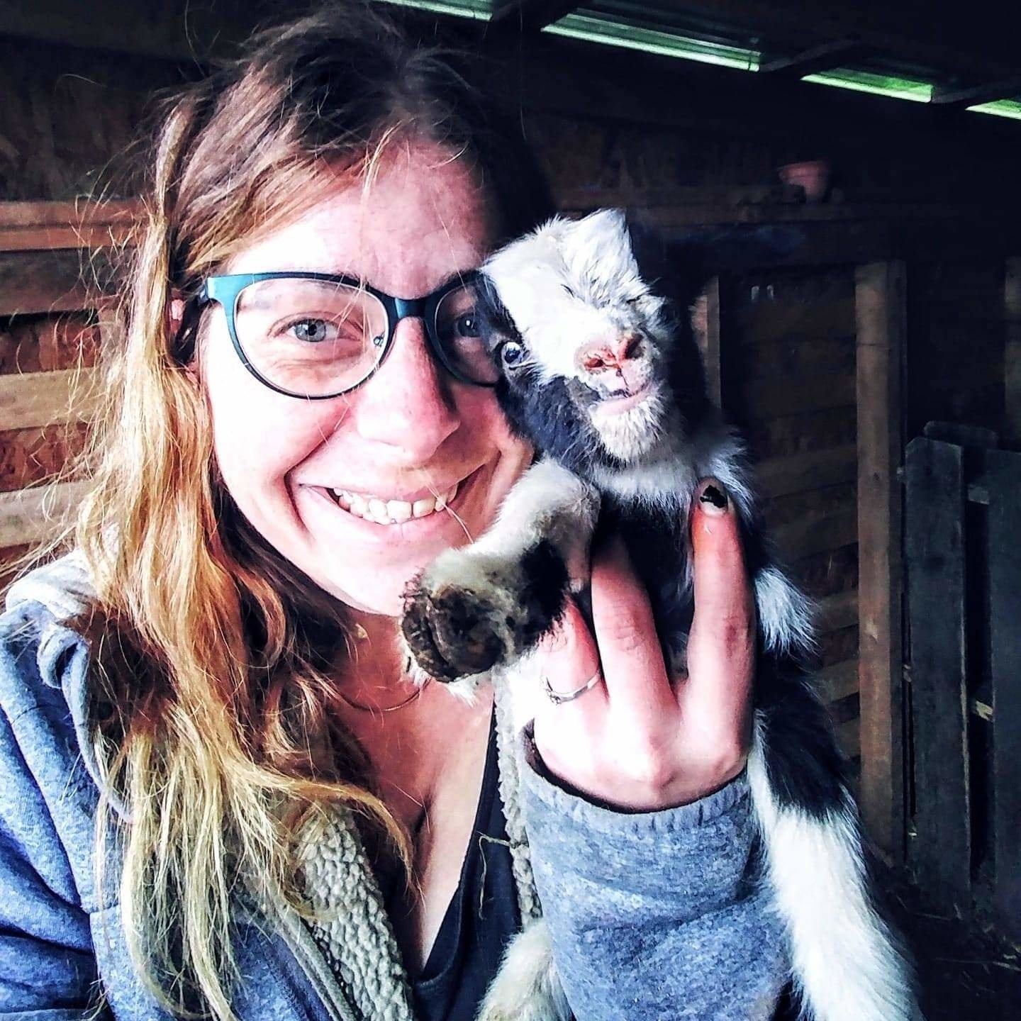 A close up of Alicia grinning at the camera while holding a tiny baby goat next to her face!