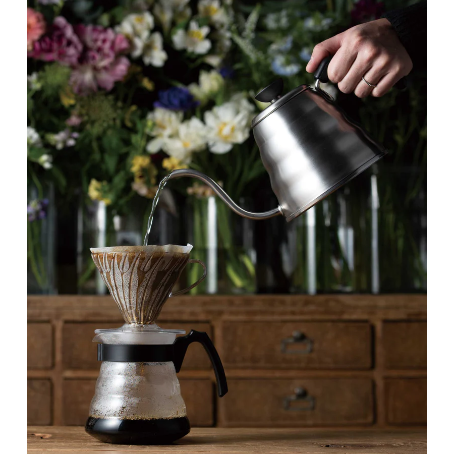 a hand holds a kettle pouring hot water into a hario v60 brewer overtop a carafe.