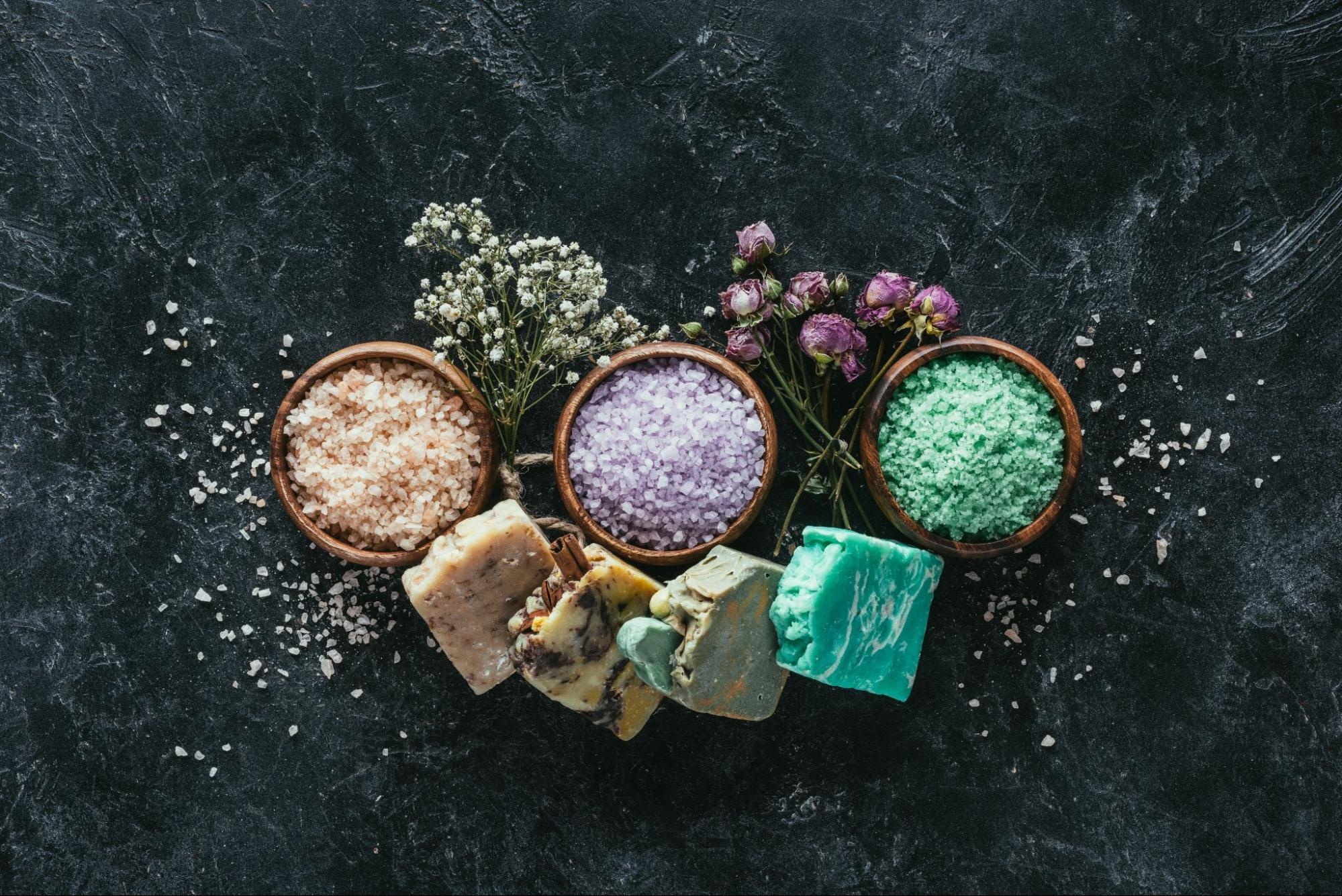 Aerial view of homemade soap, dried flowers, and sea salt in wooden bowls on a black marble surface.