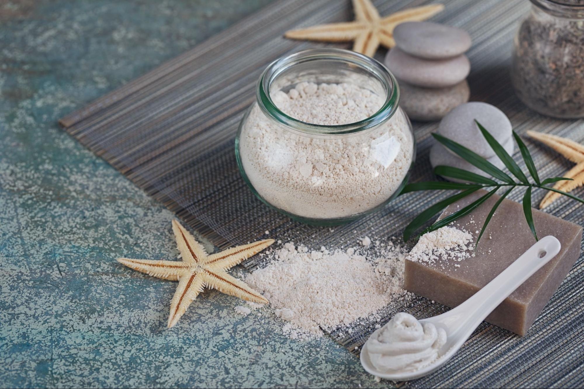 A container full of bath salts on a mat surrounded by starfish, sea stones, and a cream.