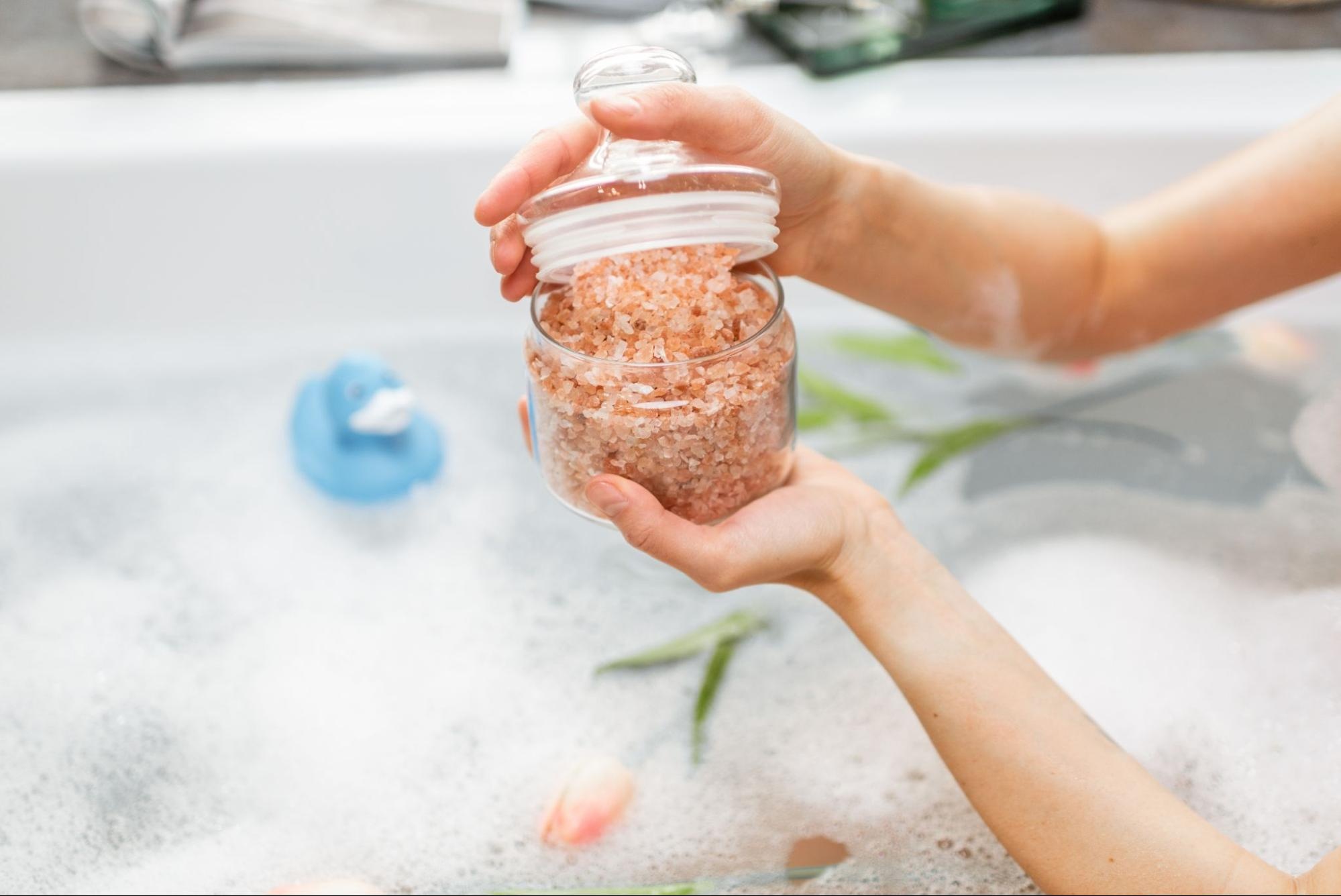 Woman holding a jar containing pink bath salts over a bathing tub.