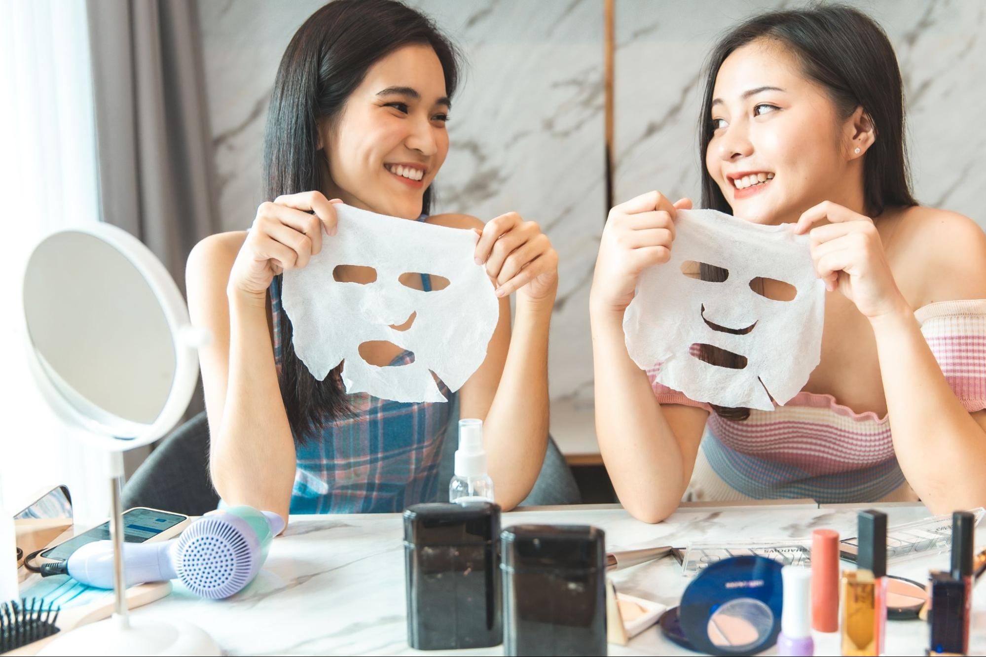 Two women in a bathroom smiling at each other while holding white sheet masks.