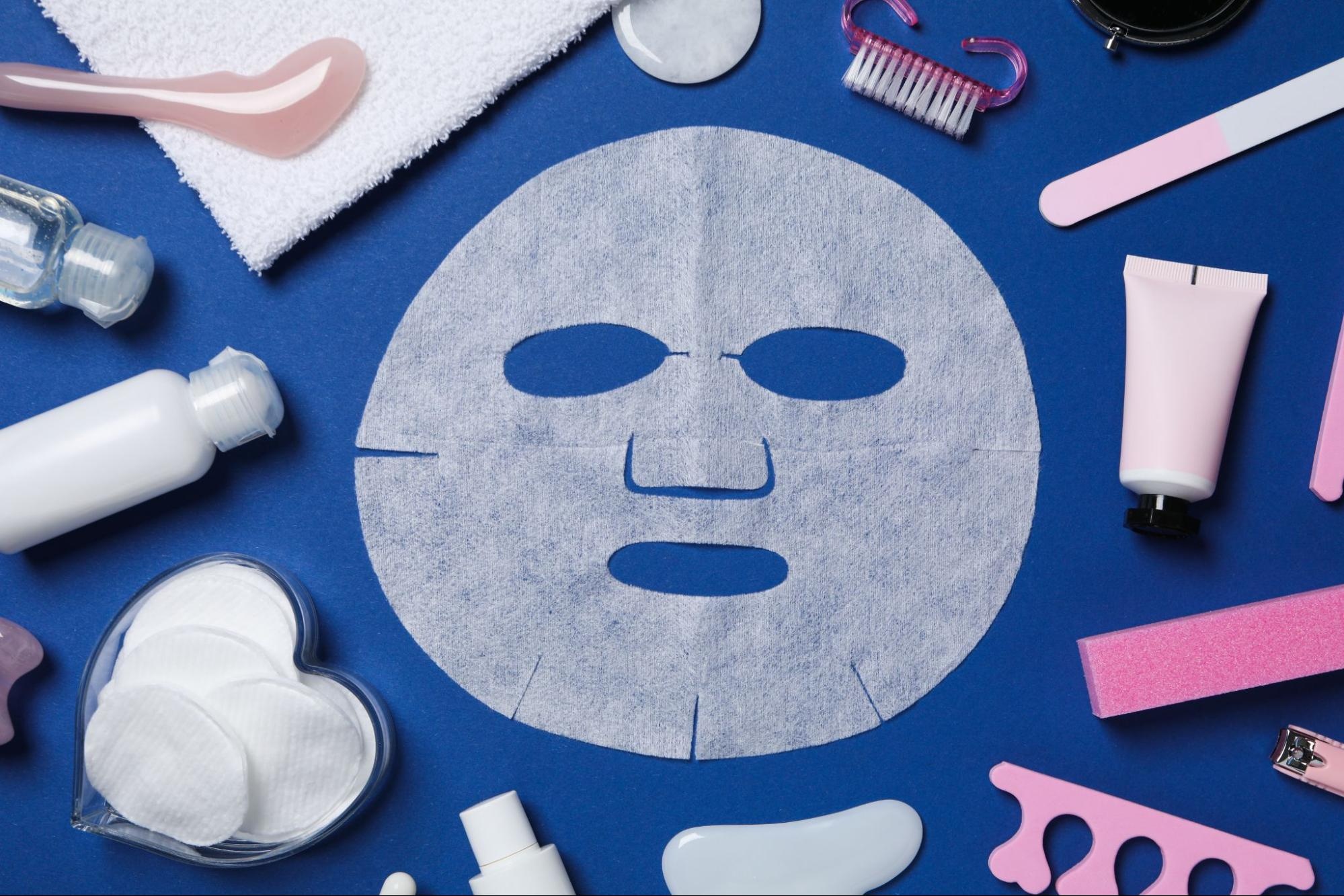 A sheet mask next to various beauty items against a blue background