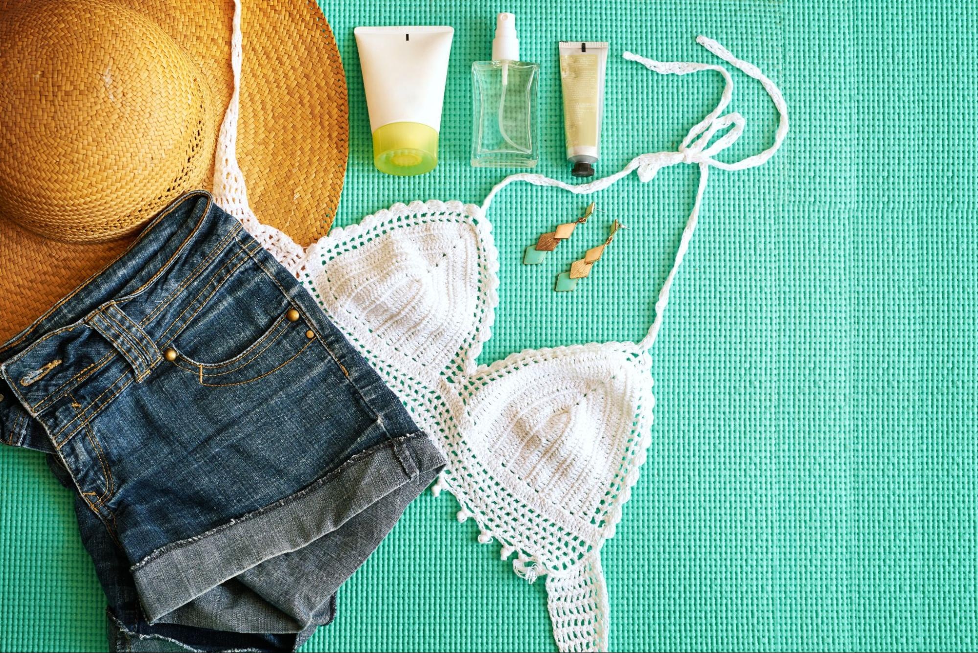 A hat, jean shorts, white tank top, earrings, and sunscreen against a turquoise background.