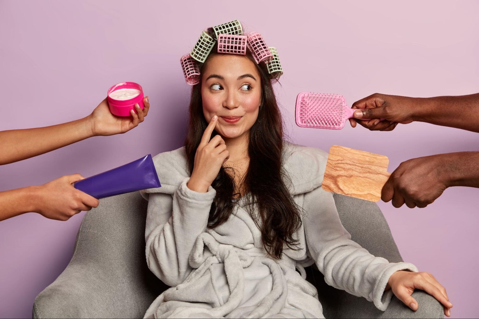 Woman sitting with hair curlers and four hands extending different self-care products to her. 