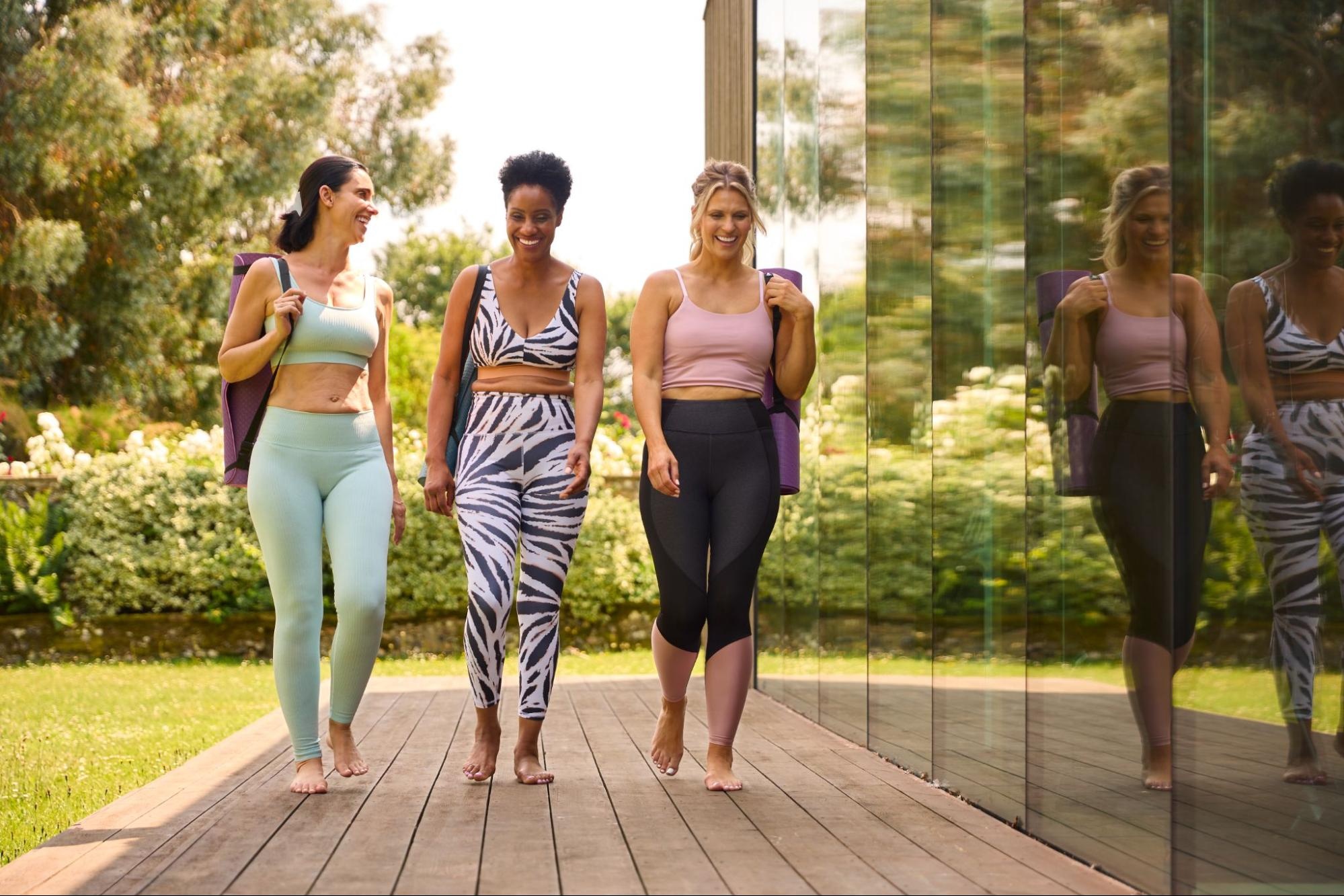Three women walking outside and wearing leggings with yoga mats slung over their shoulders. 