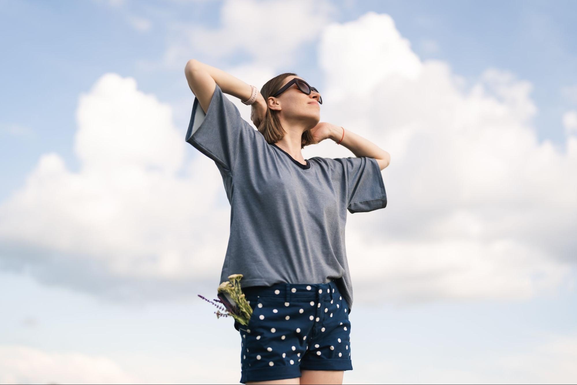 Woman in shorts and sunglasses with her arms extended with clouds in the background. 