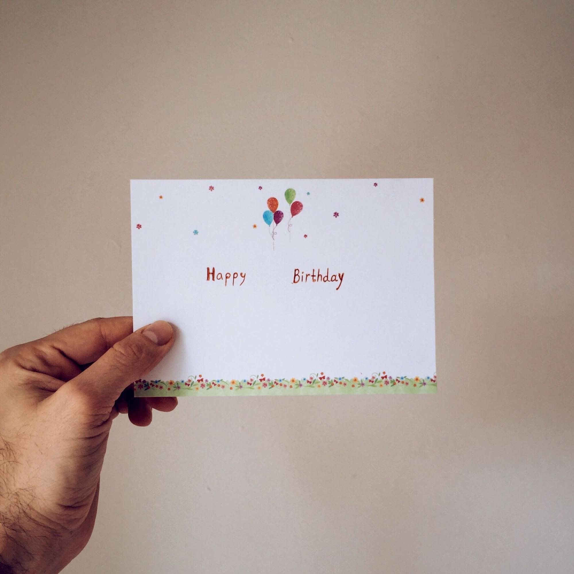A close-up view of someone's hand holding a white birthday card that says "Happy Birthday," with balloons at the top and colorful dots at the bottom.