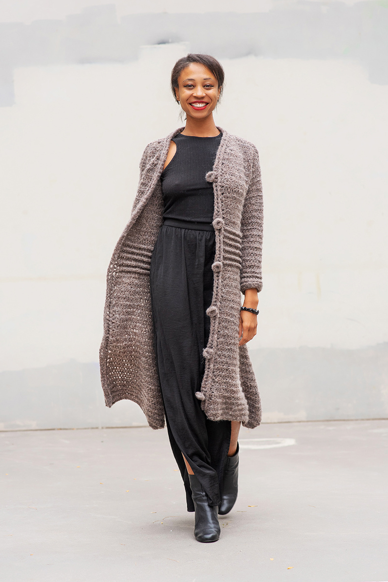 A black woman faces the camera as though she's mid-stride walking forward. She wears a calf length textured lavender crocheted coat.