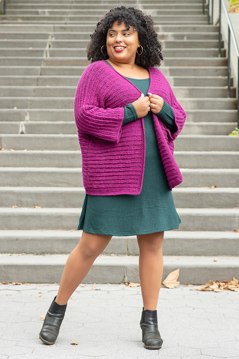 A Black woman stands in front of a staircase wearing a cozy magenta crocheted sweater. She is looking over her shoulder smiling.