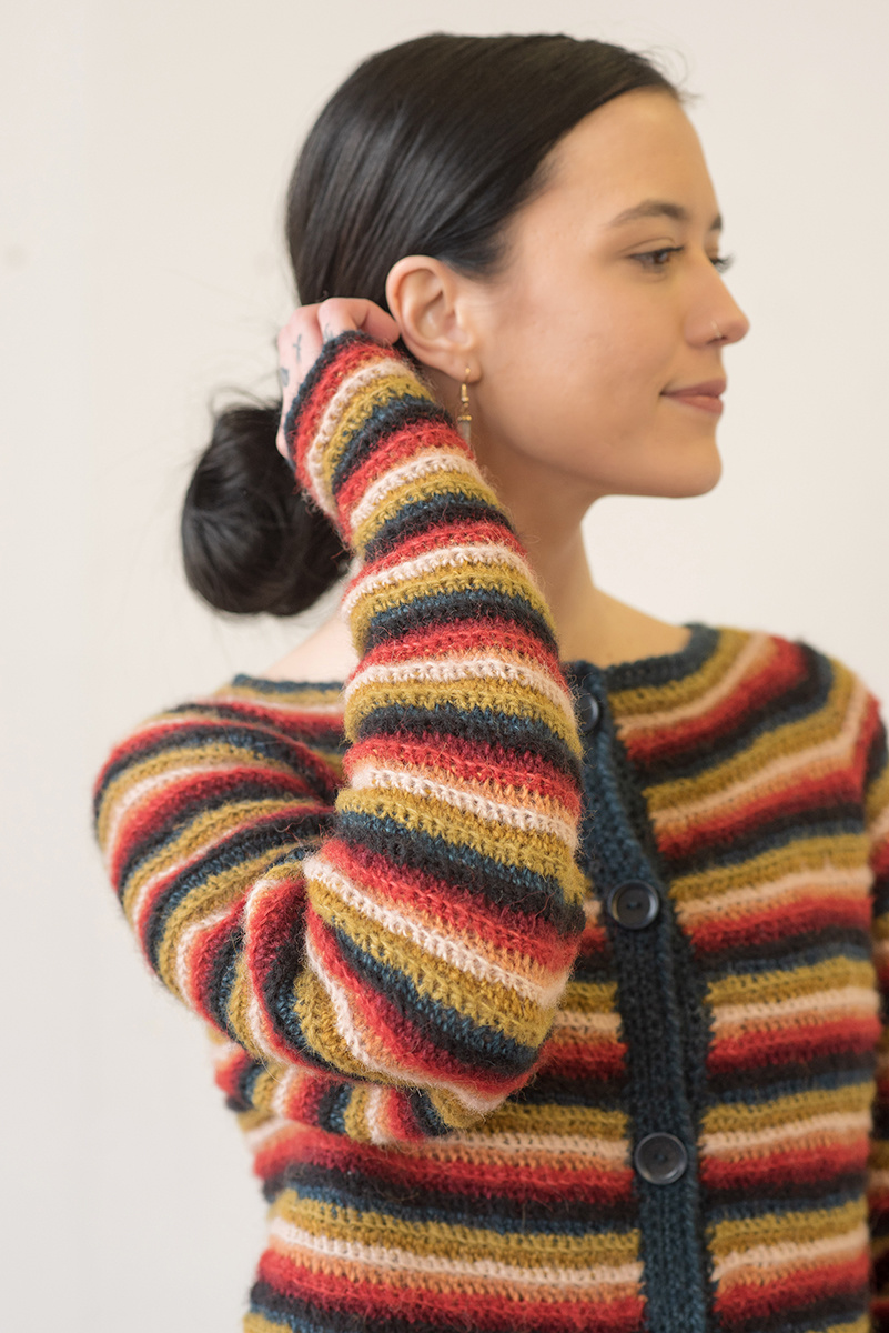 A dark-haired woman tucks her hair behind her ear. She wears a crocheted striped cardigan full of color - red, yellow, black and more.