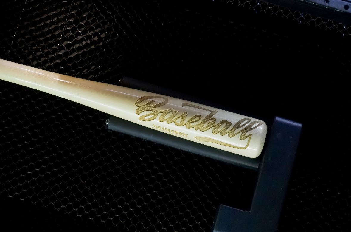 Baseball bat inside a FLUX Beambox laser cutter, held by the rotary module. The bat is engraved with the word Baseball.