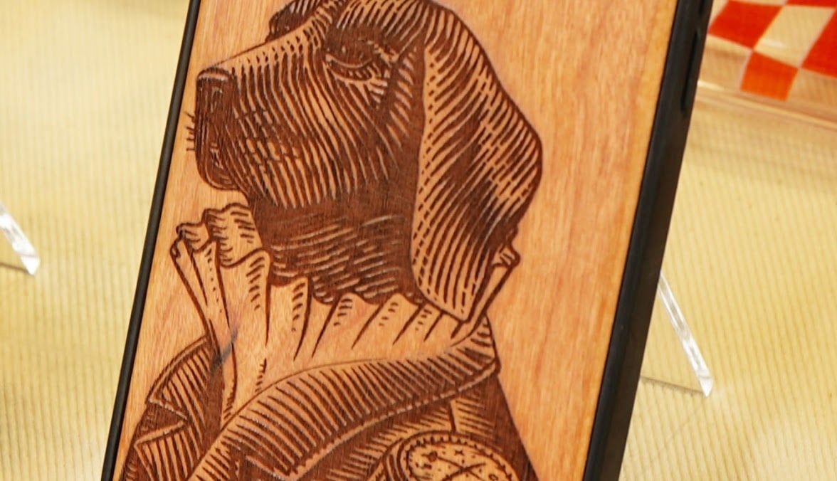 Close-up of a wooden mobile phone case with an engraving of the face of a dog in period dress in woodcut style.