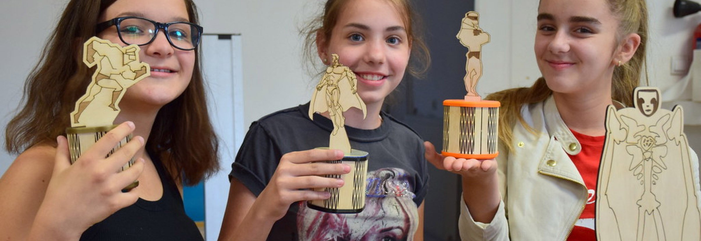 Three smiling girls holding laser cut and engraved superheroes on a round base.