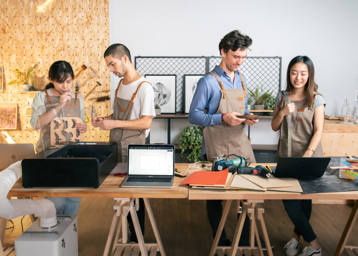 Four people standing behind a wooden table in pairs. The man and a woman on the left are looking at an open FLUX Beambox Pro. Next to it is an open laptop. The woman is holding a wooden sheet with butterflies cut out. The man and woman on the right are looking at a laptop. The man is holding some wooden disks, the woman is holding a ruler. In front of them is an array of wooden materials they can laser cut and some powertools.