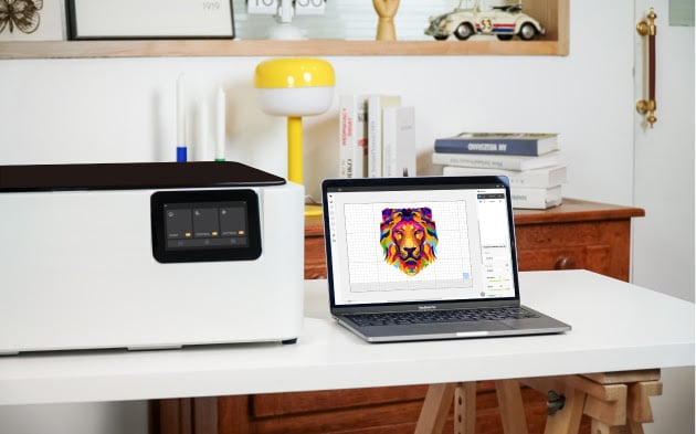 FLUX Ador sitting on a whote wooden table next to an open laptop showing Beam Studio. A file of a colorful lion's head is shown on screen.