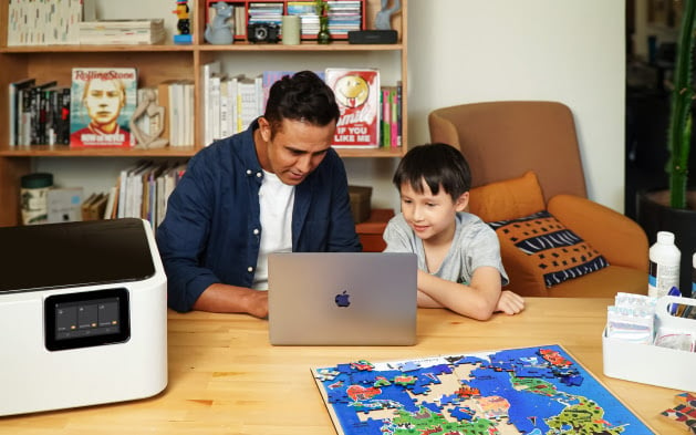 Man and young boy sitting at a table and working on a laptop in a living room setting. On the table is a FLUX Ador and a colorful wooden laser cut puzzle of a world map.