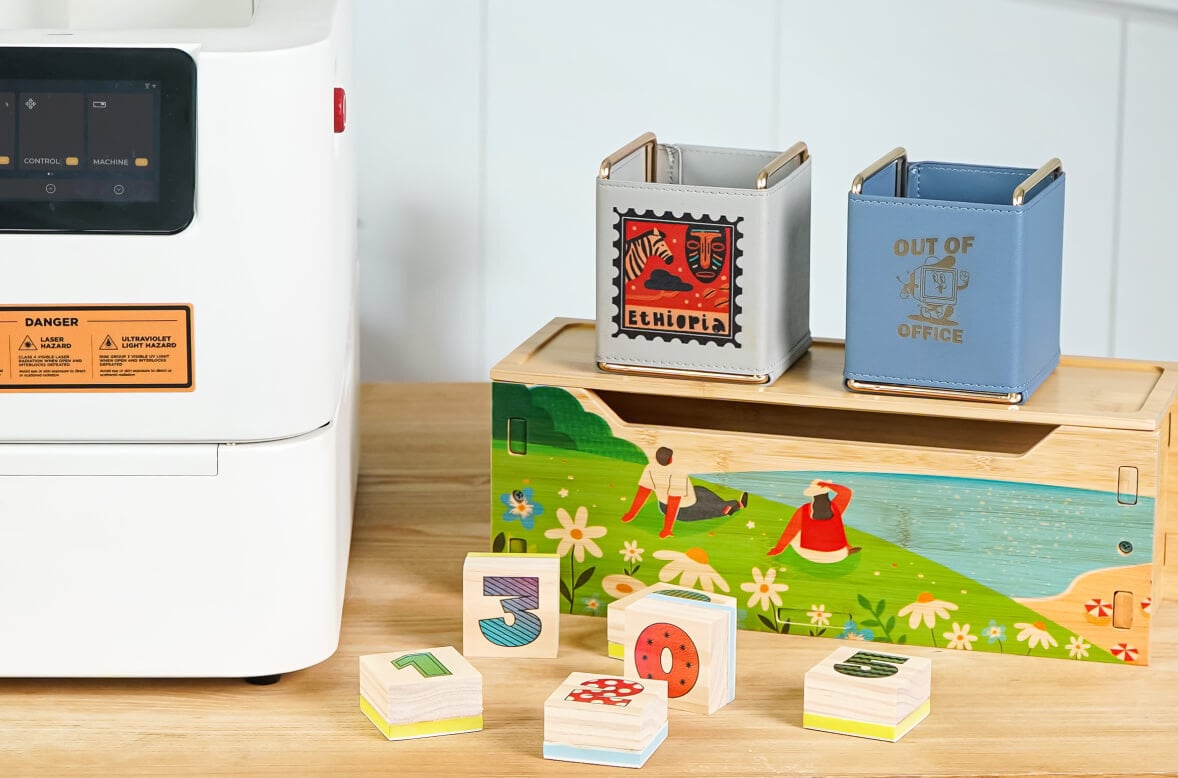 Wooden table with a partial view of a FLUX Ador with the extension base add-on on the left. On the right are six wooden stamps with numbers printed on them. Behind them is a wooden box with a landscape and two people printed on the side. Sitting on top are two cubes with a leather wraparound. One has a print of a stamp with a zebra and mask, the other has an engraved cartoon computer and the text 'Out of office'.