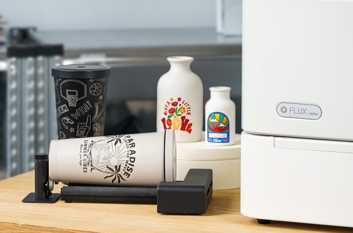 Partial view of a FLUX Ador with the extension base on a wooden table. On the left is the rotary module with a metal tumbler balancing on the rubber rollers. It has an engraved design of two palm trees, a sun and a wave and the text 'Paradise, catch the wave'. Behind it is another tumbler in black with an all-over engraving of sports themed illustrations. On its right side are two white vases with printed designs.