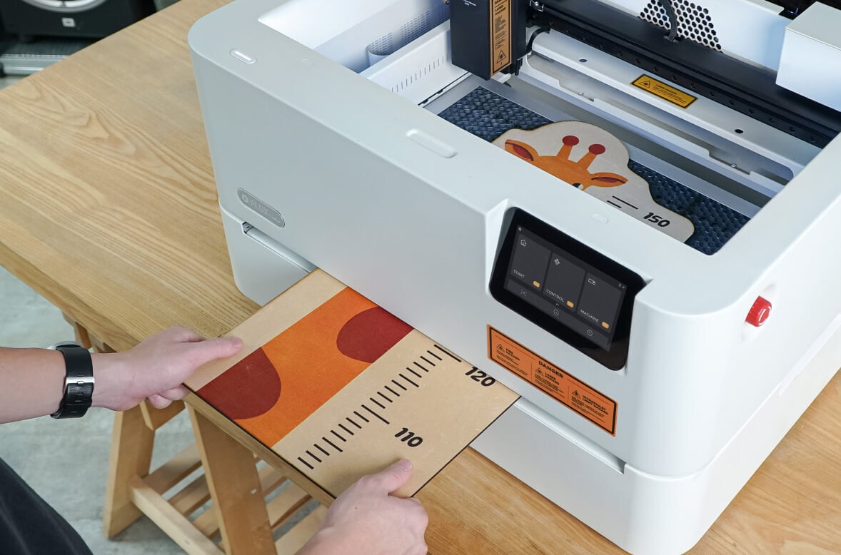 Bird's eye view of an open FLUX Ador. Two hands are feeding a large piece of wood into the extension base using the pass-through feature. It has the print of a giraffe's neck and a measuring scale.