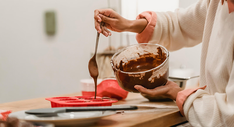 Woman holding a spoon and pouring melted chocolate in a red silicone mold.