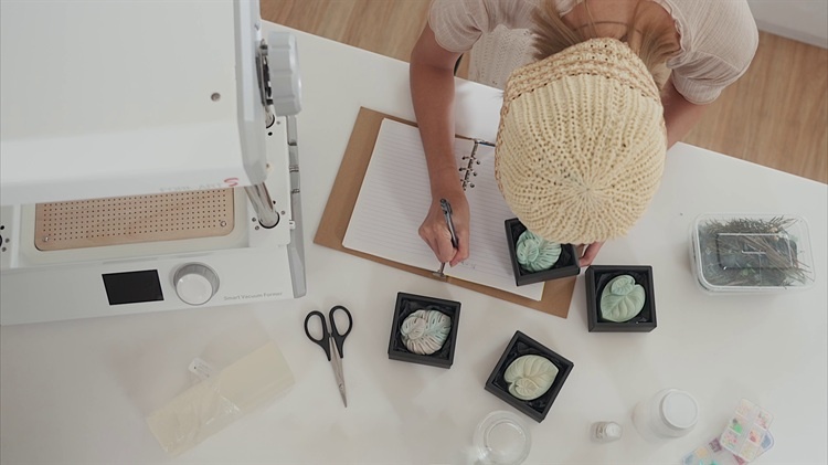 A woman taking notes on a desk filled with items she made with her MY YARD vacuum former