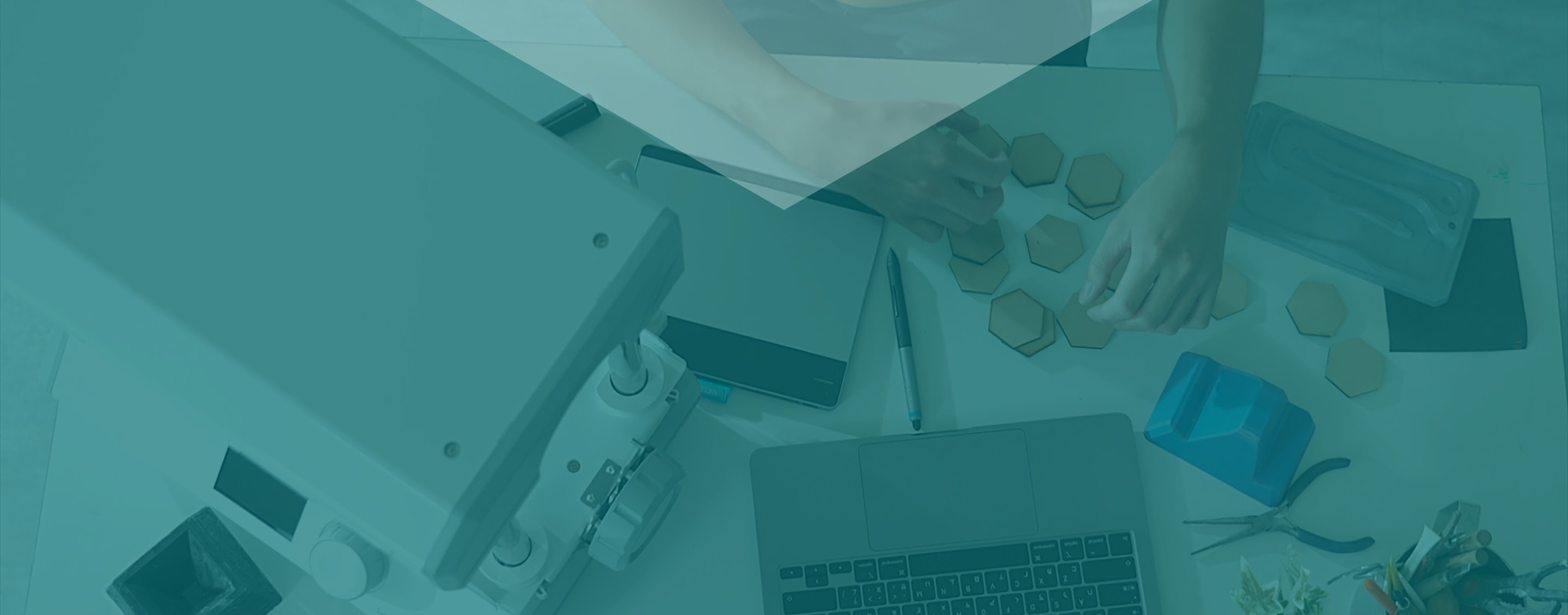 Top view of a desk. On the desk are a MY YARD vacuum former, an open laptop, blue 3D printed shape and several wooden hexagon shapes. Two hands are visible.