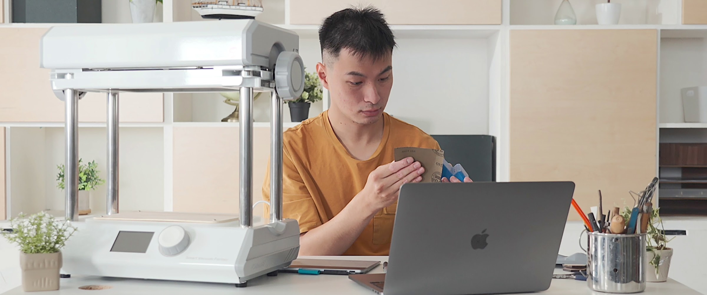 A young man in an orange shirt is sitting behind a desk sanding a 3D printed shape. On the desk are an open laptop and MY YARD vacuum former