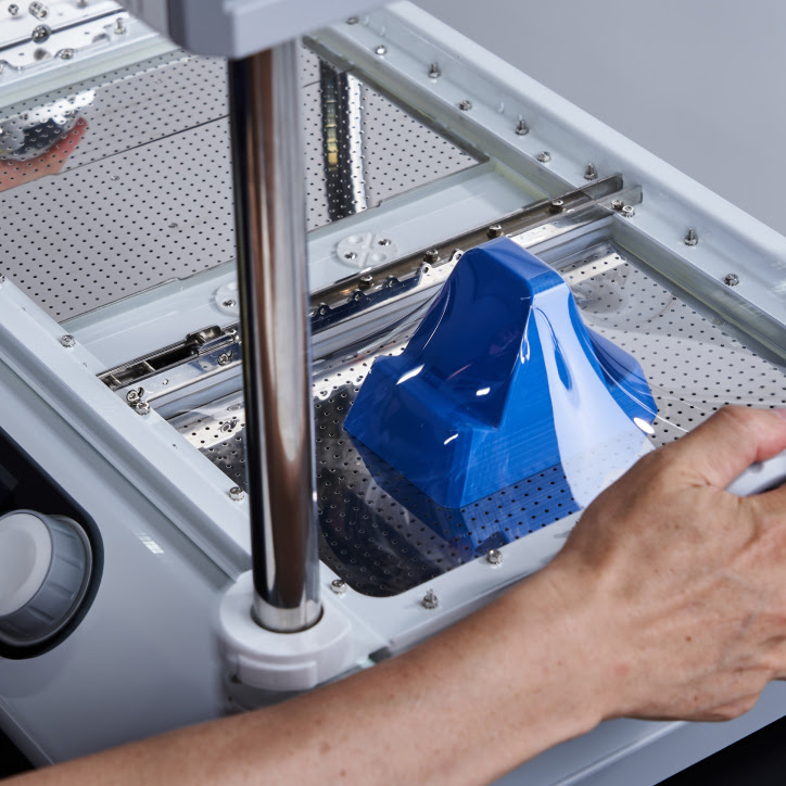 Two hands pull a clear PET sheet down on a blue 3D printed shape.