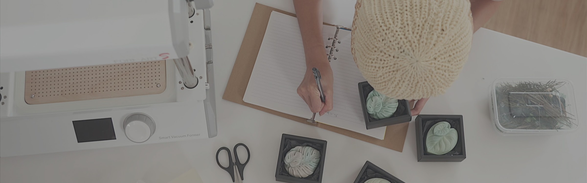 Top down view of a white desk with a MY YARD FORMART S. A woman in a beanie hat is leaning over the desk making notes in a notebook. She is holding a black box with a piece of soap in the shape of a leaf. Several similar boxes are also on the desk.